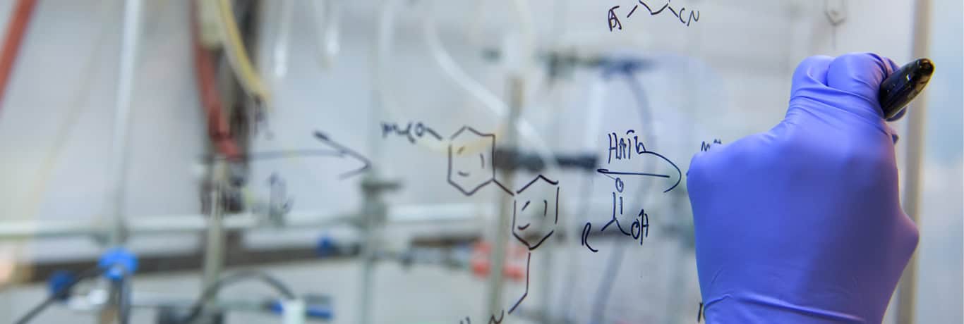 A scientist wearing a blue glove writes chemical formulas on a glass board in a laboratory.