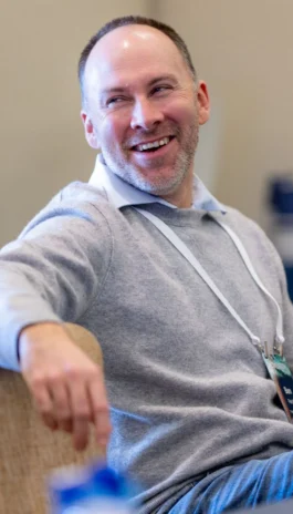 A man with a light beard and short hair, wearing a grey sweater and conference lanyard, sits and smiles while looking to the side.