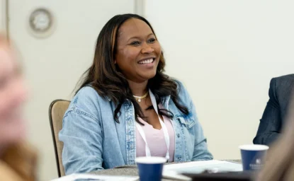 A woman wearing a denim jacket and conference badge smiles while sitting at a table during a meeting.